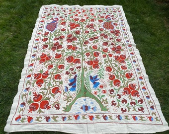 Puede incluir: Un tapiz de algodón blanco con un diseño cosido a mano de un árbol con granadas rojas, hojas verdes y pájaros azules. El borde está decorado con un patrón repetido de granadas, hojas y pájaros.