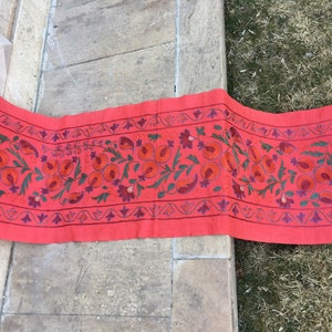 May include: A long, red runner with embroidered floral designs in shades of red, green, and purple. The runner features a repeating pattern of stylized flowers and pomegranates.
