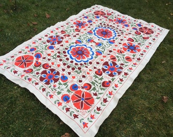 Puede incluir: Un textil rectangular de color blanco roto con intrincados motivos florales bordados a mano en tonos vibrantes de rojo, granate intenso y azul brillante. El diseño incluye grandes flores circulares centrales rodeadas de formas de granada.