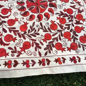 May include: White fabric tablecloth with red and brown embroidered pomegranate designs. The tablecloth has a border of small red and brown floral motifs.