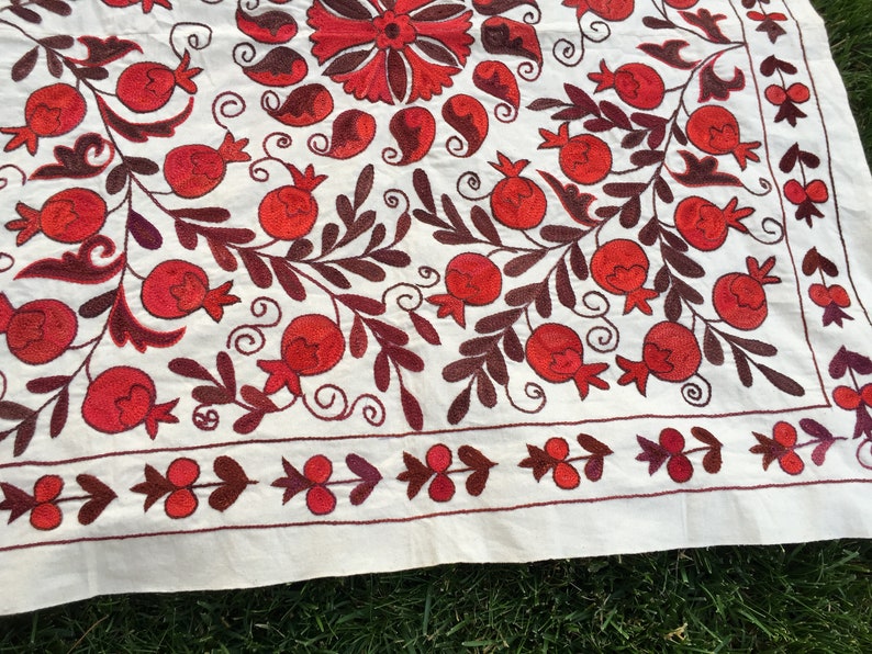 May include: White fabric tablecloth with red and brown embroidered pomegranate designs. The tablecloth has a border of small red and brown floral motifs.