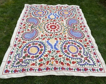 Puede incluir: Un mantel de tela de algodón blanco con bordados intrincados en tonos de rojo, azul, verde y amarillo. El diseño presenta granadas estilizadas, patrones de paisley y motivos florales.