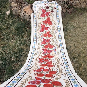 May include: White table runner with embroidered red pomegranates, green leaves, and blue birds on a white background. The runner has a green and blue border.