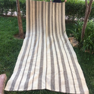 May include: A long, white and brown striped rug with a woven texture. The rug is folded in half and laid out on a grassy lawn.
