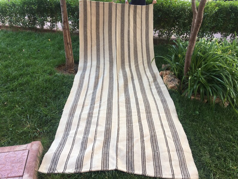 May include: A long, white and brown striped rug with a woven texture. The rug is folded in half and laid out on a grassy lawn.