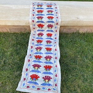 Puede incluir: Un camino de mesa blanco con un patrón floral en rojo, azul y verde. Las flores están bordadas en la tela.