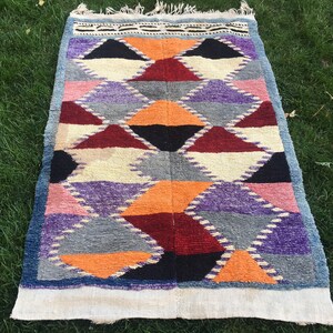 May include: A colorful geometric rug with a white border. The rug features a pattern of triangles in shades of blue, purple, orange, red, and black.