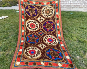 Puede incluir: Un tapiz grande, rectangular, de color marrón y rojo, bordado con un patrón repetitivo de motivos florales coloridos. El tapiz está hecho de una tela suave y texturizada y tiene un borde con flecos.