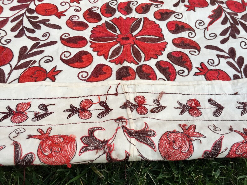 May include: A white fabric tablecloth with red and brown embroidery of floral and paisley designs. The tablecloth has a border of smaller floral designs.