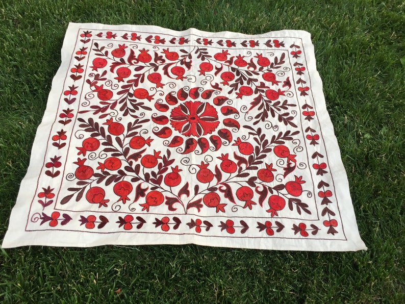 May include: A white linen square embroidered with a red and brown floral design. The design features pomegranates, leaves, and swirls.