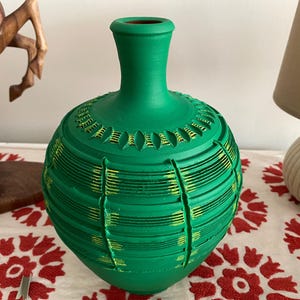 Op de afbeelding: Een groene keramische vaas met een ronde body en een smalle hals. De vaas heeft horizontale en verticale getextureerde lijnen met gele accenten. De vaas wordt tentoongesteld op een oppervlak met bloemmotief.