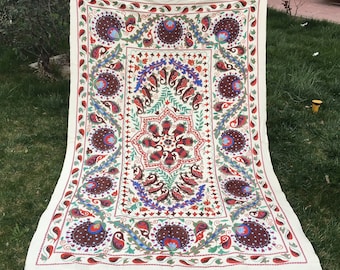 Puede incluir: Un tapiz rectangular de tela con fondo blanco roto y motivos florales y de cachemira bordados a mano en tonos rojo oscuro, azul, verde y naranja, dispuestos en un patrón simétrico con un medallón central.