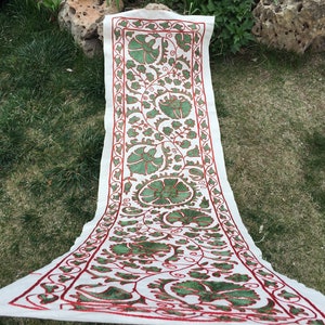 May include: A white fabric runner with a red and green floral embroidery design. The runner is folded in half and lying on a green grassy surface.