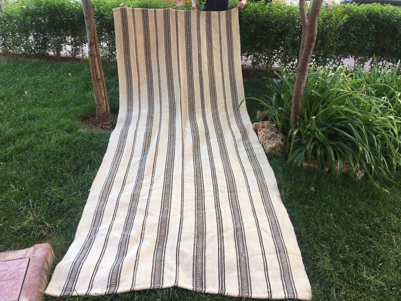 May include: A long, white and brown striped rug with a woven texture. The rug is laid out on a grassy lawn.