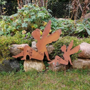 Può includere: Due sagome di fate in metallo arrugginito, una più grande dell'altra, sono posizionate in un ambiente da giardino. Le fate sono raffigurate con le ali e in pose sedute.