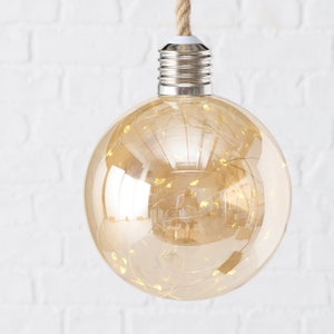 Può includere: Una grande lampadina rotonda in vetro trasparente con una base in metallo argentato e una stringa di luci LED bianco caldo all'interno. La lampadina è appesa a una corda bianca.