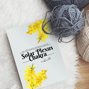 May include: A gray and white affirmation release journal with the title "Solar Plexus Chakra" and the text "by @Roseth1" on the cover. The journal is lying on a white fuzzy surface with two balls of gray yarn.