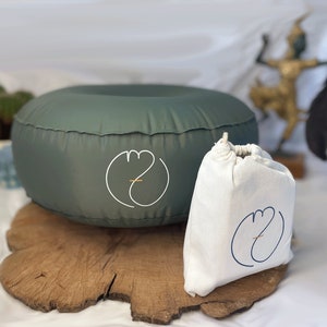 Può includere: Un cuscino da meditazione gonfiabile verde salvia e un sacchetto con coulisse abbinato. Entrambi gli articoli presentano un logo bianco con un accento dorato. Il cuscino è circolare e poggia su una lastra di legno, suggerendo relax e consapevolezza.