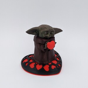 May include: A brown 3D printed figurine of a character holding a red heart, standing on a black heart-shaped base with red hearts cut out.