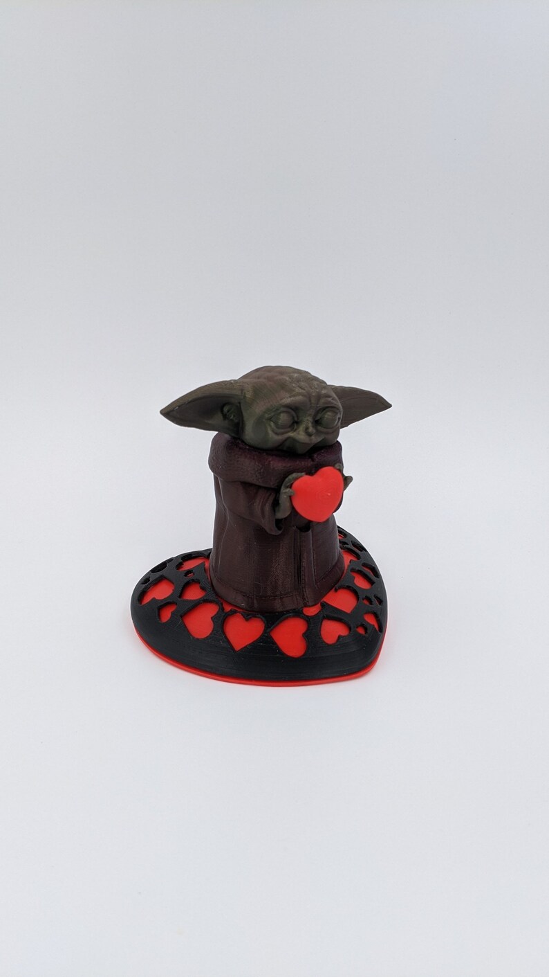 May include: A brown 3D printed figurine of a character holding a red heart, standing on a black heart-shaped base with red hearts cut out.