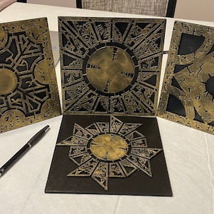 May include: Four black square wooden panels with gold metallic designs. Each panel features a different intricate pattern with a circular center. The panels are arranged in a square shape on a white surface.