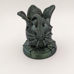 May include: A dark green 3D printed figurine of an alien creature emerging from an egg. The creature has a long, segmented tail and is curled up inside the egg. The egg is cracked open and has a textured surface.