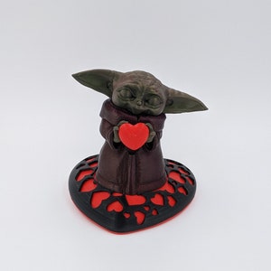 May include: A green and brown figurine of a character holding a red heart. The figurine is standing on a black heart-shaped base with red hearts cut out.