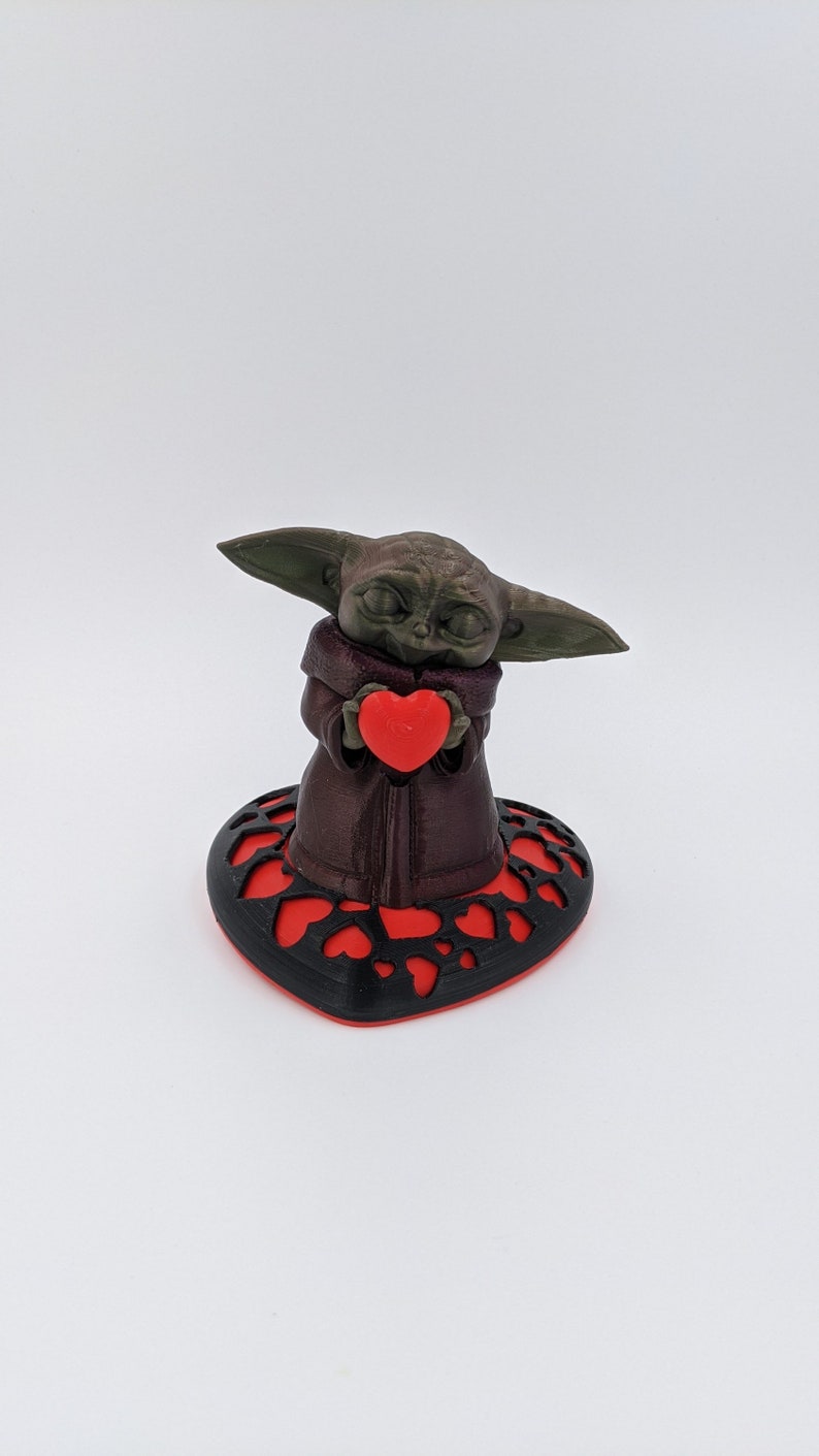 May include: A green and brown figurine of a character holding a red heart. The figurine is standing on a black heart-shaped base with red hearts cut out.