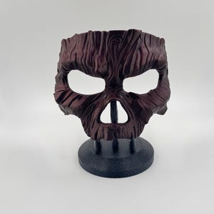 May include: A dark burgundy mask with a wood grain texture, featuring eye and mouth openings. The mask is displayed on a black stand with a circular base. The mask has a carved, organic appearance.