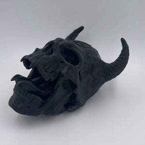 May include: A black, 3D printed skull with horns and an open mouth, resembling a demon. The skull is detailed with teeth and a tongue.