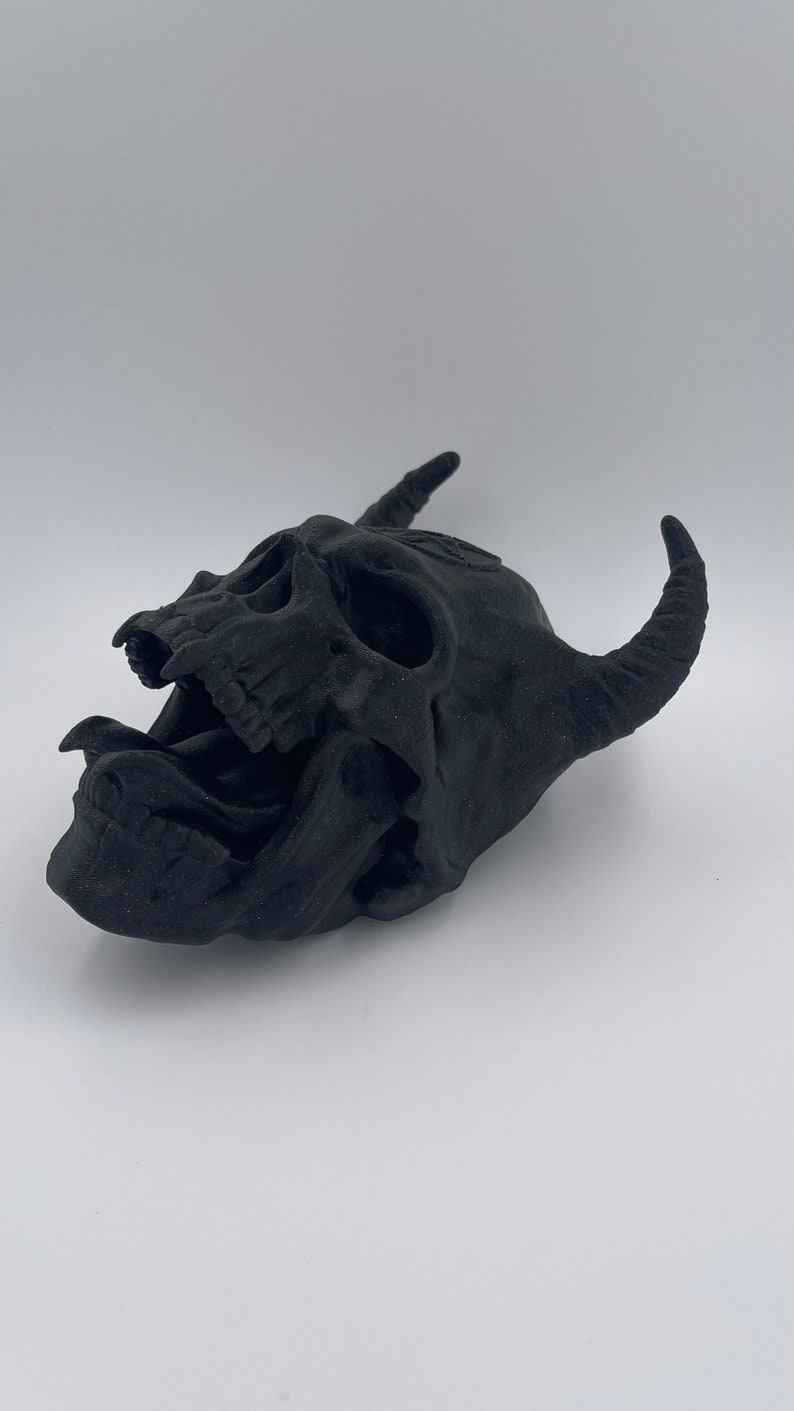 May include: A black, 3D printed skull with horns and an open mouth, resembling a demon. The skull is detailed with teeth and a tongue.