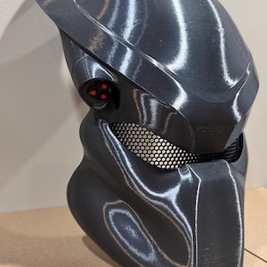 May include: A black plastic Predator mask with a mesh eye piece and red glowing eyes.