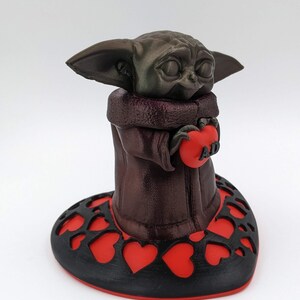 May include: A brown 3D printed figurine of a character holding a red heart with the word "AD" on it. The figurine is standing on a black heart-shaped base with red hearts cut out.