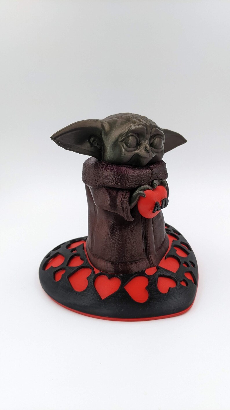 May include: A brown 3D printed figurine of a character holding a red heart with the word "AD" on it. The figurine is standing on a black heart-shaped base with red hearts cut out.