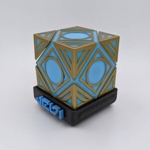 May include: A gold and blue geometric cube with a black base. The base has the word "Jedi" printed on it in blue.