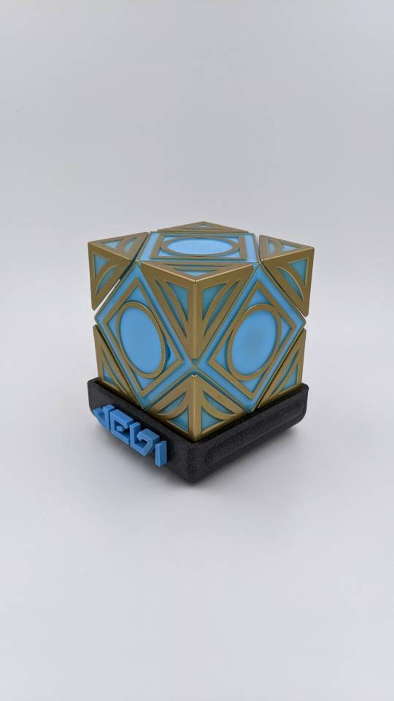 May include: A gold and blue geometric cube with a black base. The base has the word "Jedi" printed on it in blue.