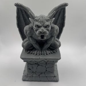 Puede incluir: Estatua de gárgola gris con alas extendidas, posada sobre un pedestal texturizado. La escultura presenta rasgos faciales detallados y una expresión amenazante, mostrando un tema arquitectónico gótico.