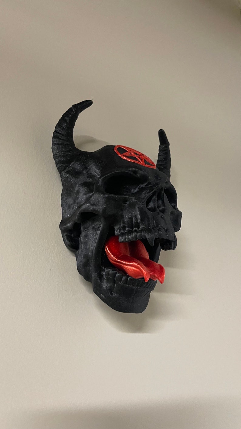 May include: A black 3D printed skull with horns and a red pentagram on the forehead. The skull has a large, red tongue sticking out.