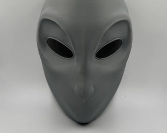 May include: A gray alien mask with large, oval eye openings and a smooth, rounded design. The mask is symmetrical and appears to be made of a solid material. The overall shape is elongated, with a pointed chin and a prominent nose.