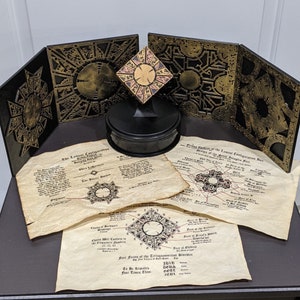 May include: A black wooden box with intricate gold designs and a smaller gold box with a similar design on top. The box is surrounded by several pieces of paper with text and diagrams. The text includes the words "The Lamesi Configuration" and "Four Forces of the Tetragrammaton Signature".