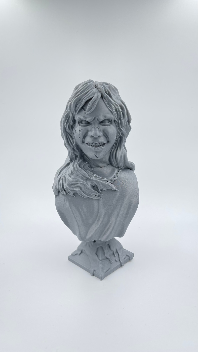 May include: A gray 3D printed bust of a female figure with a menacing expression. The figure has long hair and is wearing a simple garment. The bust is mounted on a square base.