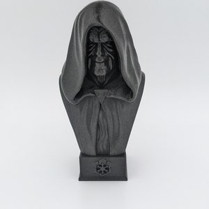 May include: A black 3D printed bust of Darth Maul, a character from the Star Wars franchise. The bust depicts Maul with his signature red and black face paint, horns, and hooded robe. The bust is sitting on a black base with the Star Wars logo.