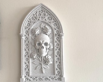 May include: A gray gothic-style wall plaque featuring a skull and roses design. The plaque has an arched top and is decorated with intricate floral and scrollwork details. The skull is centered, with roses surrounding it.