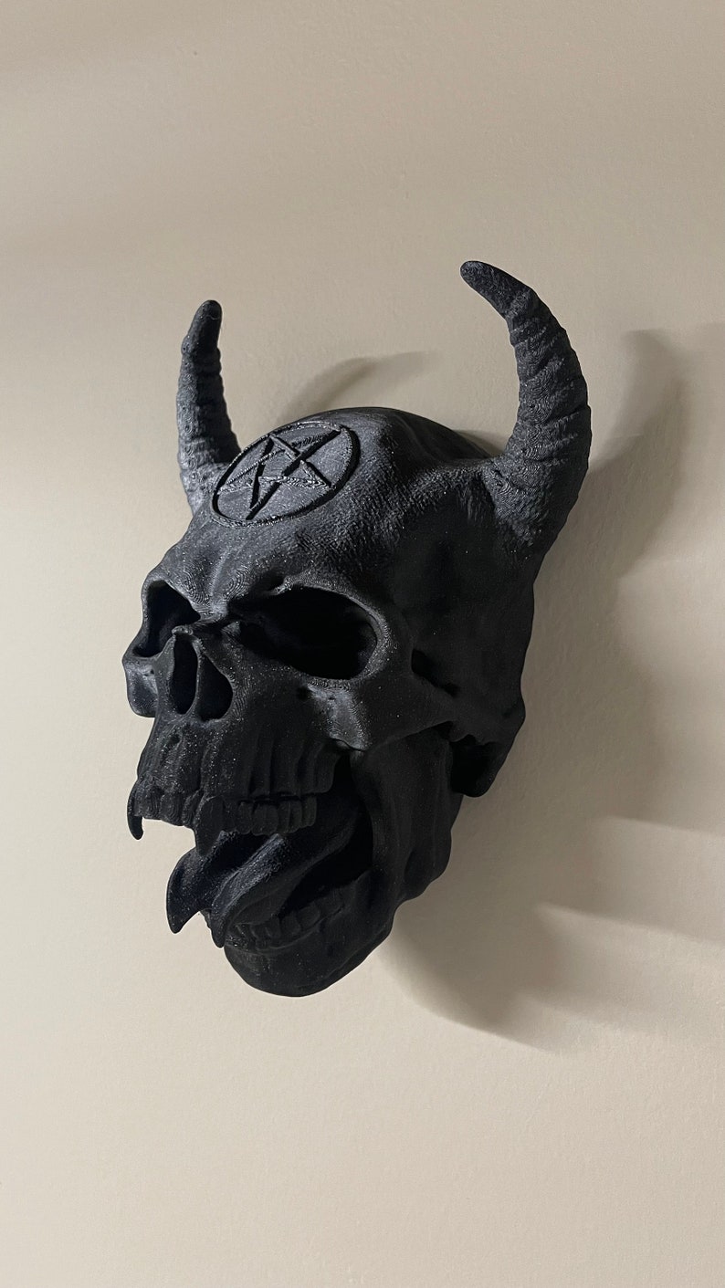 May include: A black 3D printed skull with horns and a pentagram on the forehead. The skull has a detailed design with teeth and a tongue sticking out.