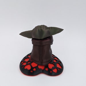 May include: A 3D printed figurine of a character from the Star Wars franchise, with a green hat and brown robe, standing on a red and black heart-shaped base.