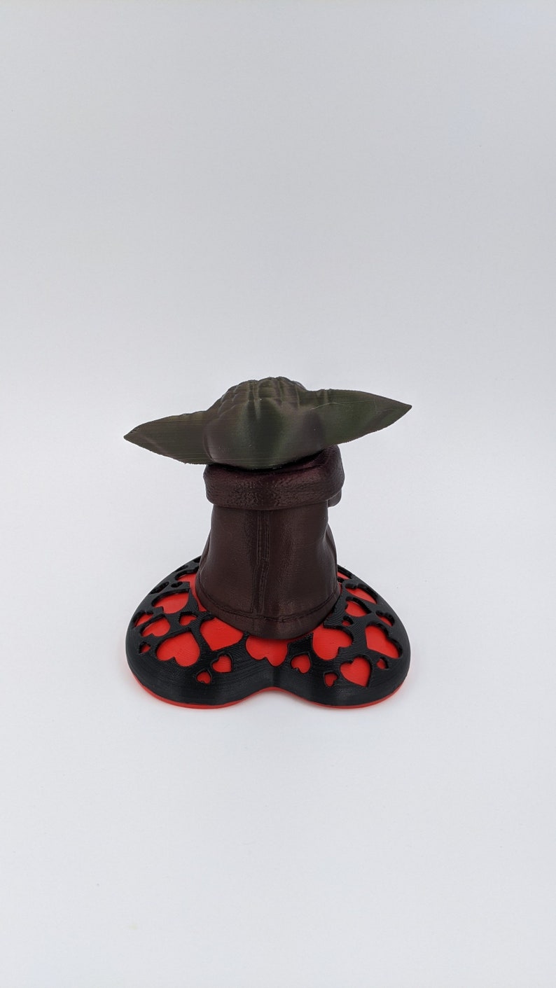May include: A 3D printed figurine of a character from the Star Wars franchise, with a green hat and brown robe, standing on a red and black heart-shaped base.