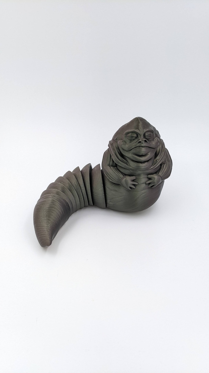 Puede incluir: Una figurita de color gris oscuro impresa en 3D de una criatura ficticia con una cabeza grande y una cola segmentada. La criatura tiene una cabeza grande y redonda con una boca ancha y los ojos cerrados. La cola est&aacute; segmentada y enroscada alrededor del cuerpo de la criatura.