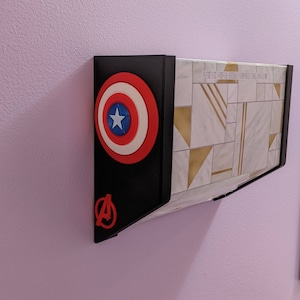 May include: A black wall mount with a red, white, and blue Captain America shield and a red Avengers logo. The mount is attached to a white wall with a geometric pattern.