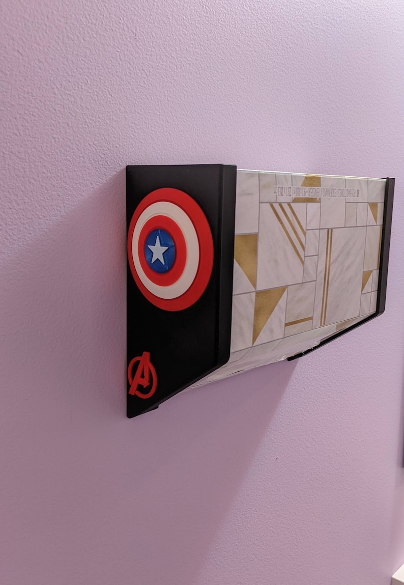 May include: A black wall mount with a red, white, and blue Captain America shield and a red Avengers logo. The mount is attached to a white wall with a geometric pattern.