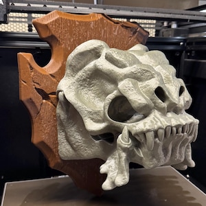 May include: A 3D-printed, light gray monster skull mounted on a brown, wooden-style plaque. The skull features sharp teeth and detailed bone texture. The plaque has a rustic, weathered appearance.